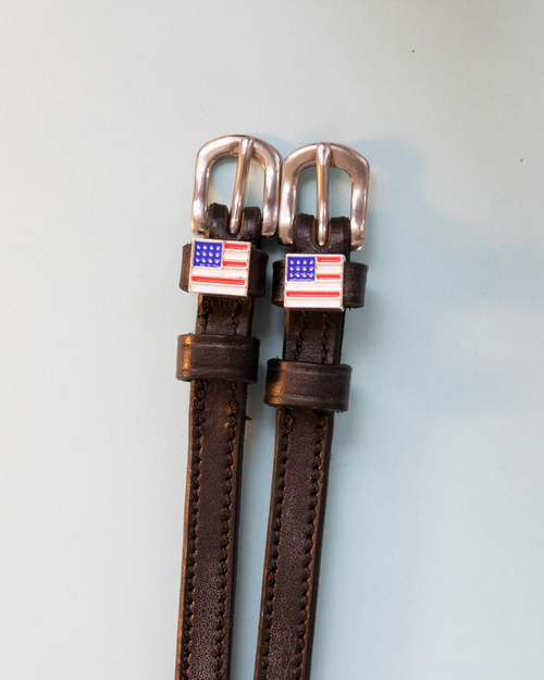 Brown leather ManeJane spur straps with American flag charms and rose-gold buckles