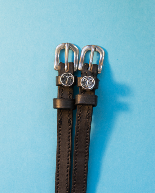 Black leather ManeJane spur straps with round Aries zodiac charms on a light blue background
