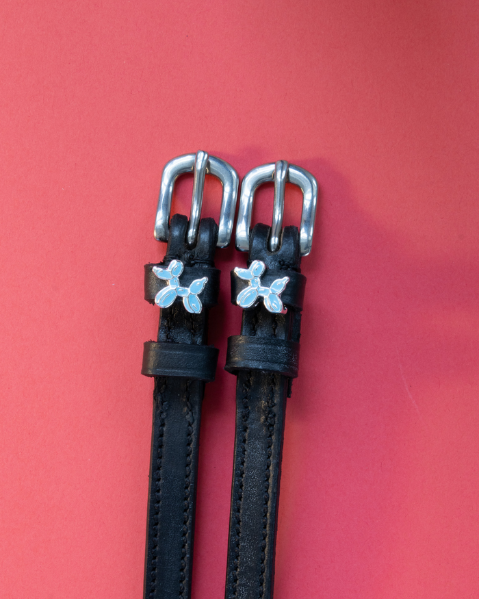 Black leather spur straps with blue balloon dog charms on a coral red background
