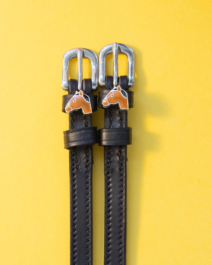 Black leather ManeJane spur straps with tan horse-head charms on a yellow background