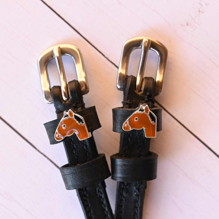 Close-up of tan horse-head charms and silver buckles on black spur straps, pink planks
