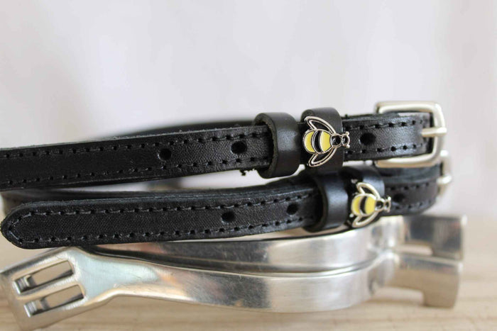 Side view of black bee spur straps resting on a silver spur, showing buckle and charm