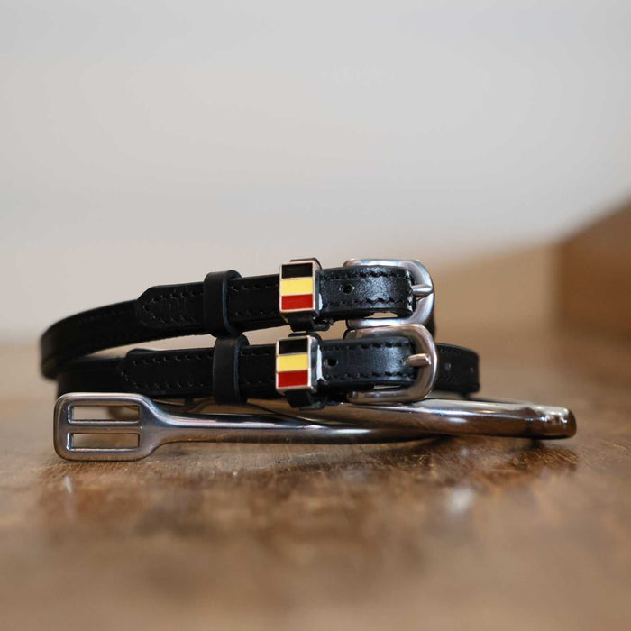 Black leather Belgium flag spur straps stacked on a silver spur on wood