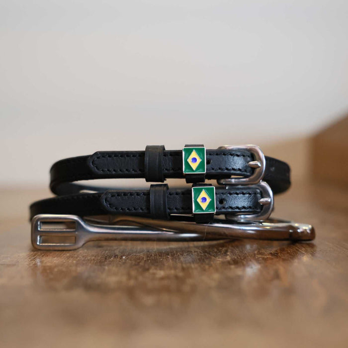 Black leather Brazil flag spur straps coiled beside a stirrup on a wooden surface