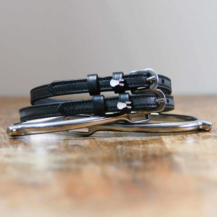 Stacked black Bunny Hop spur straps with silver buckles on wood beside a stirrup iron