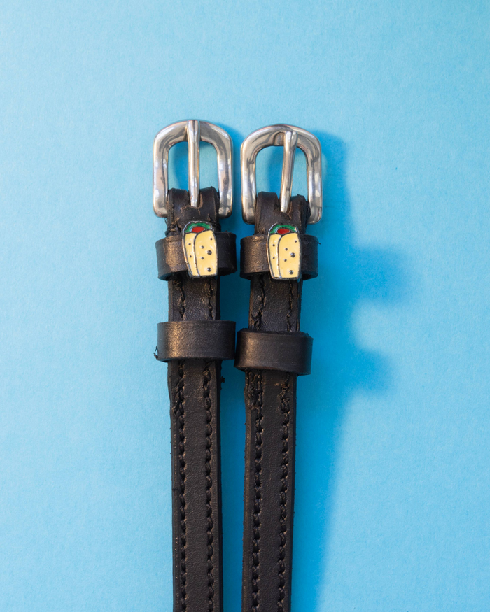 Pair of black leather spur straps with burrito charms flat-laid on a light blue background