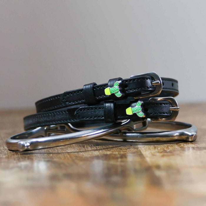 Cactus charm spur straps coiled beside a stirrup on a wooden tabletop