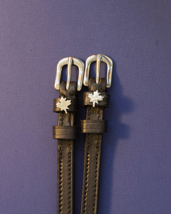 Black leather ManeJane spur straps with silver buckles and maple leaf charms on a purple background