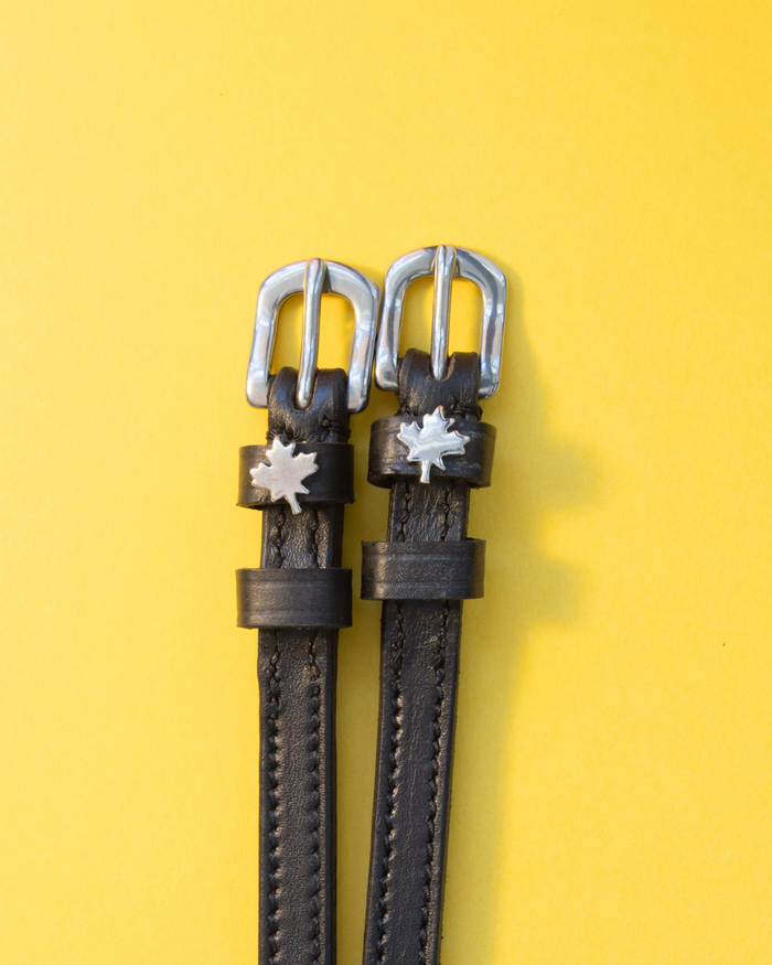 Black leather maple leaf ManeJane spur straps with silver buckles on a yellow background