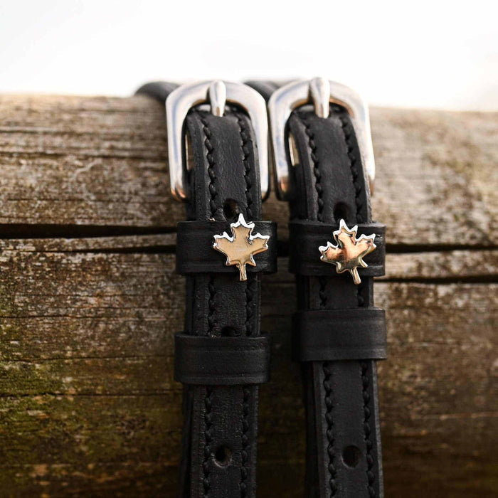 Close-up of silver and gold maple leaf charms on black leather spur straps against weathered wood
