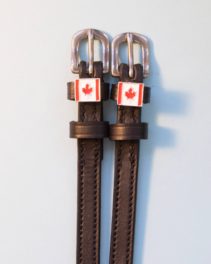 Brown leather ManeJane spur straps with silver buckles and Canadian flag charms on grey