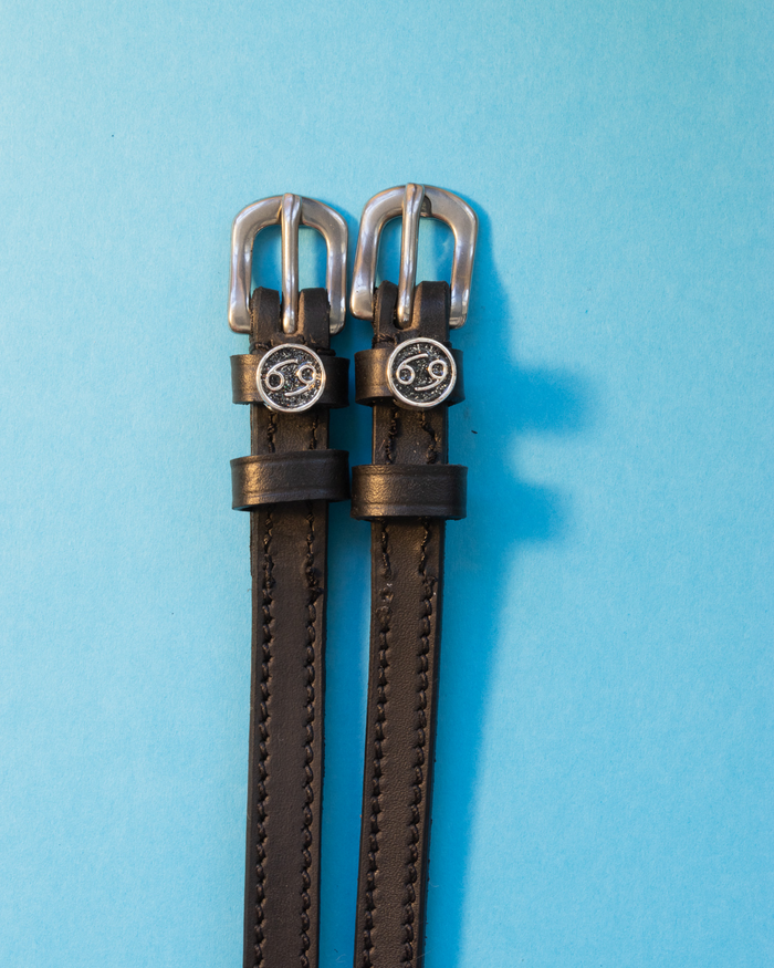 Black leather ManeJane spur straps with round Cancer zodiac charms on a light blue background
