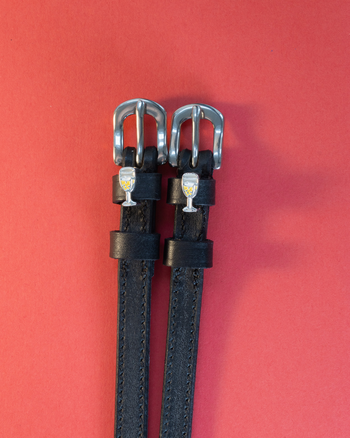 Black leather ManeJane spur straps with champagne-glass charms on a coral background