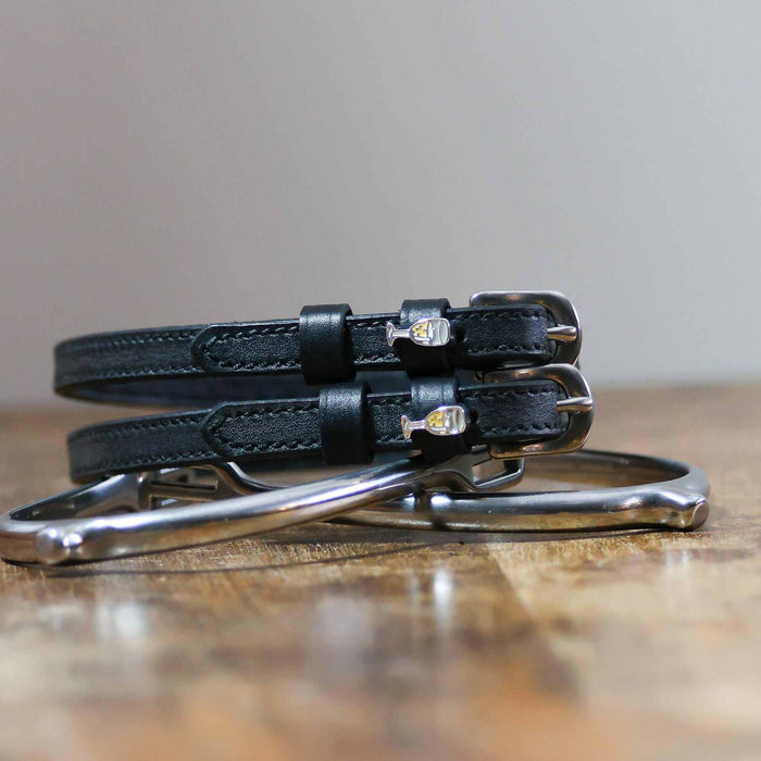 Black leather champagne spur straps coiled on wood beside a metal stirrup