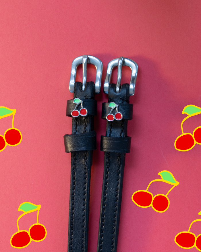 Pair of black leather ManeJane spur straps with silver buckles and red cherry charms on a red backdrop