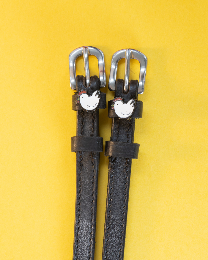 Black leather ManeJane spur straps with silver buckles and white hen charms on a yellow background