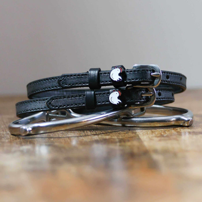 Black leather chicken spur straps coiled beside a silver stirrup on a wooden surface