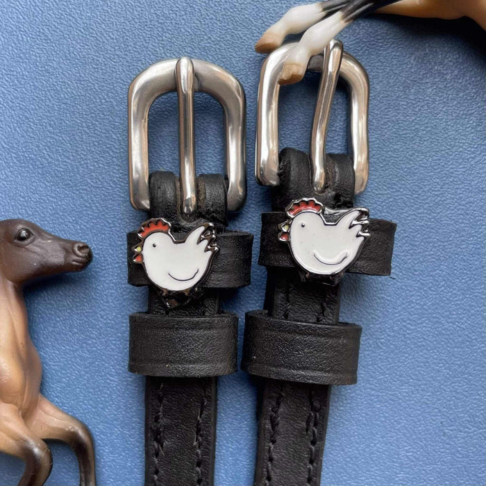 White hen charm spur straps with silver buckles beside a horse figurine on blue
