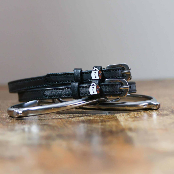 Black leather coffee-mug spur straps coiled on wood beside a stirrup