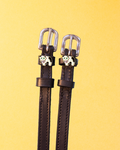 Dark brown leather ManeJane cow spur straps with silver buckles and enamel cow charms on a yellow background