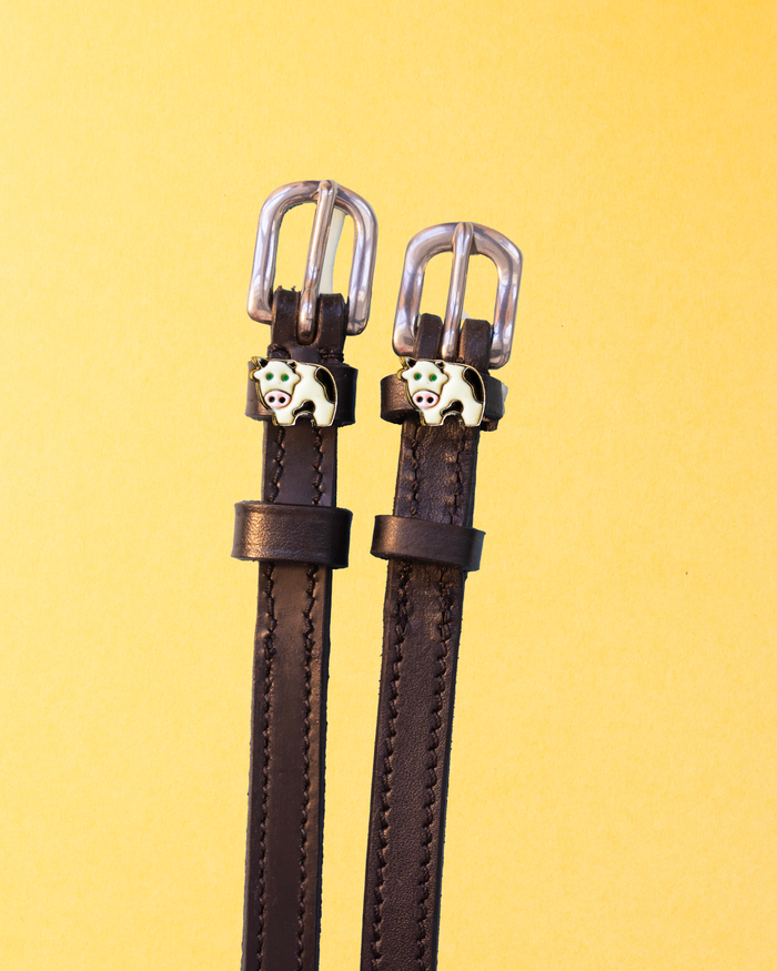 Dark brown leather ManeJane cow spur straps with silver buckles and enamel cow charms on a yellow background