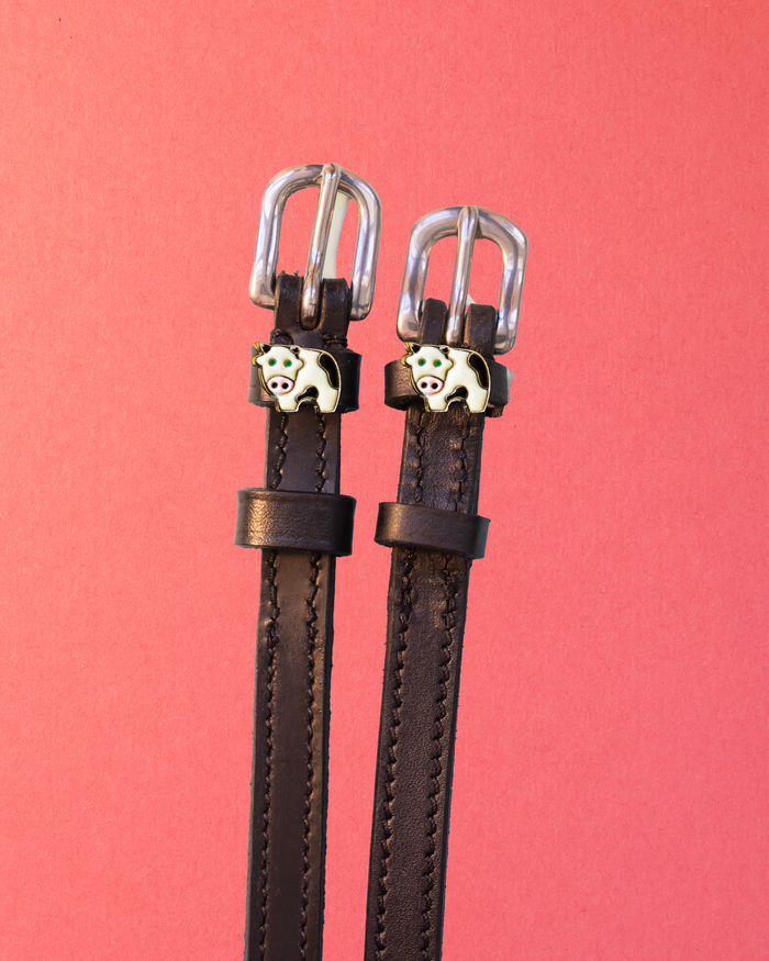 Pair of black leather ManeJane cow spur straps with enamel cow charms on a coral background