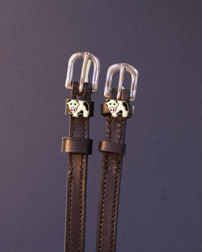 Brown leather ManeJane cow spur straps with silver buckles and cow charms on a dark slate background