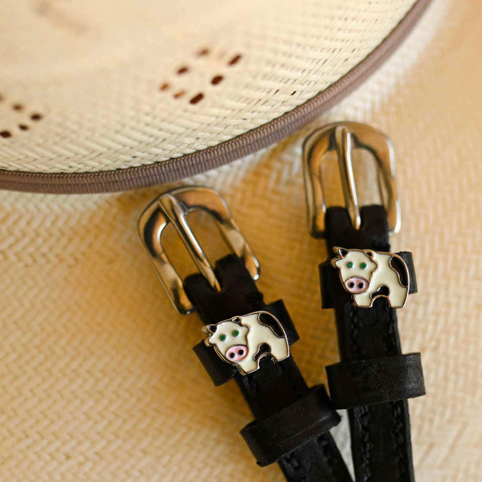 Black leather cow spur straps beside a straw sun hat, ManeJane enamel cow charm close-up