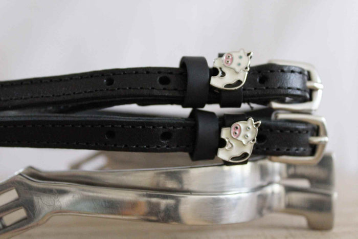 Side view of black leather ManeJane cow spur straps with cow charms resting on a silver stirrup iron