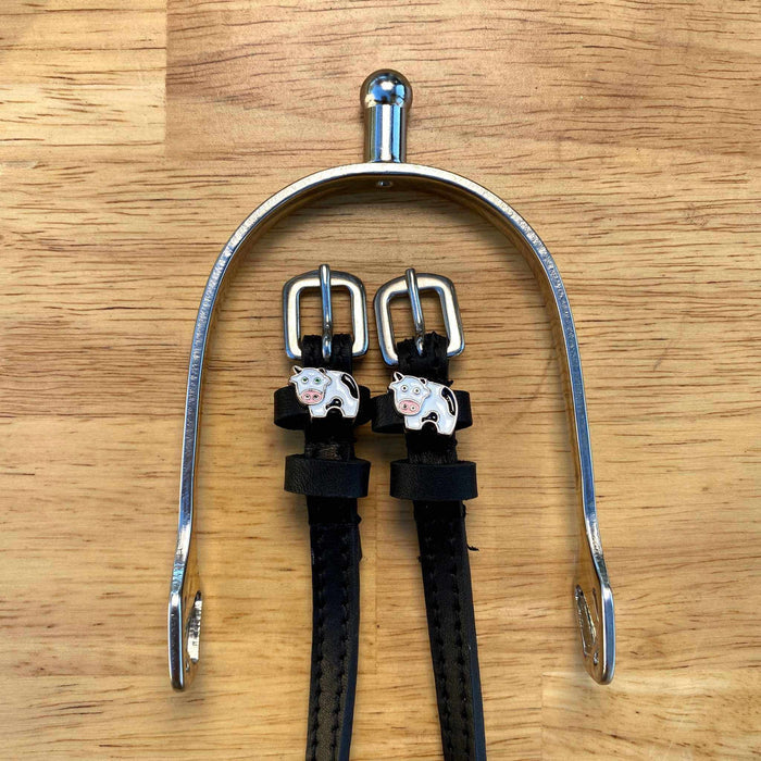 Black leather cow spur straps laid over a silver spur on a wooden surface, ManeJane cow charms