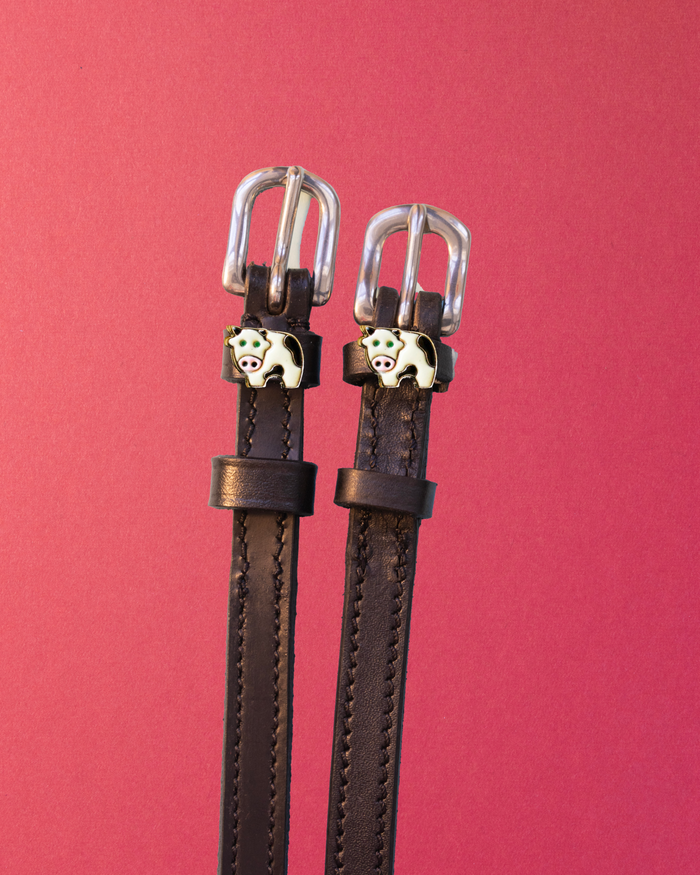 Dark brown ManeJane cow spur straps with enamel cow charms on a rich red background