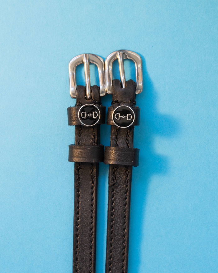 Black leather ManeJane spur straps with round snaffle-bit charms on light blue