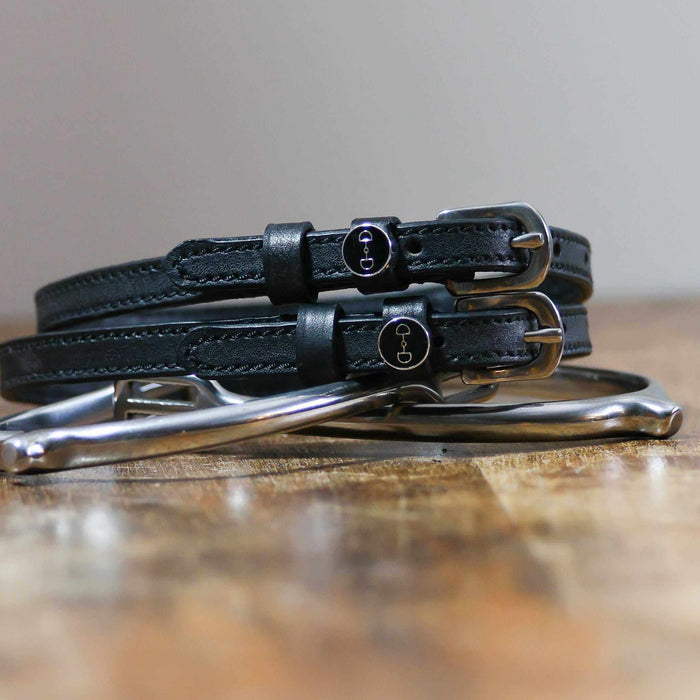 Black leather D-ring spur straps coiled on wood beside a stirrup