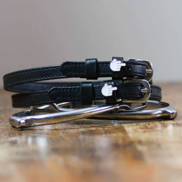 Stacked black Da Bird spur straps with silver buckles on wood beside a stirrup iron, ManeJane