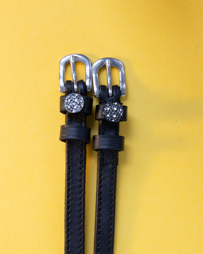Black leather disco ball charm spur straps with silver buckles on a yellow background