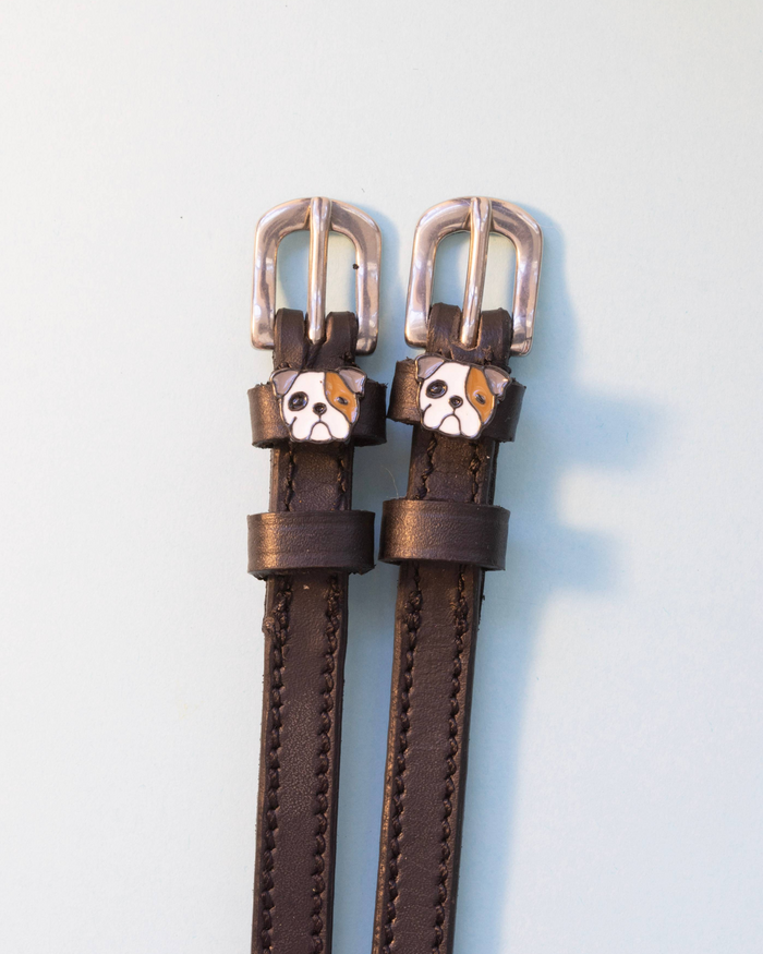 Dark brown leather ManeJane spur straps with bulldog head charms on a pale background