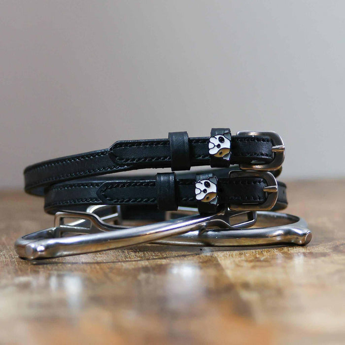 Black leather bulldog spur straps coiled on wood beside a silver bit