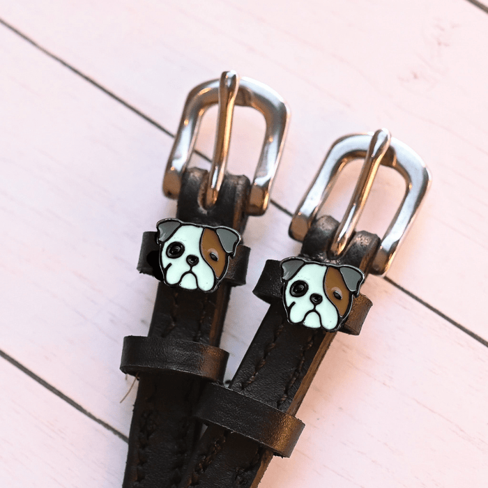 Bulldog head charms on dark brown leather spur straps over pale pink wood planks