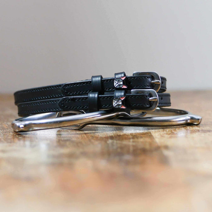 Black leather Frenchie spur straps coiled beside a stirrup on a wooden surface
