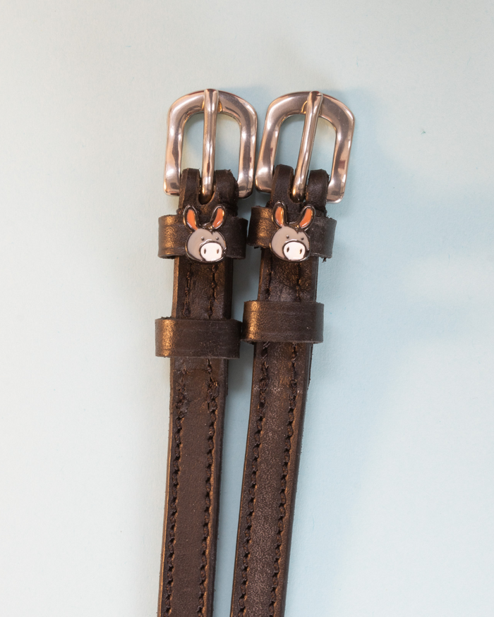 Brown leather ManeJane donkey spur straps with rose-gold buckles on pale blue