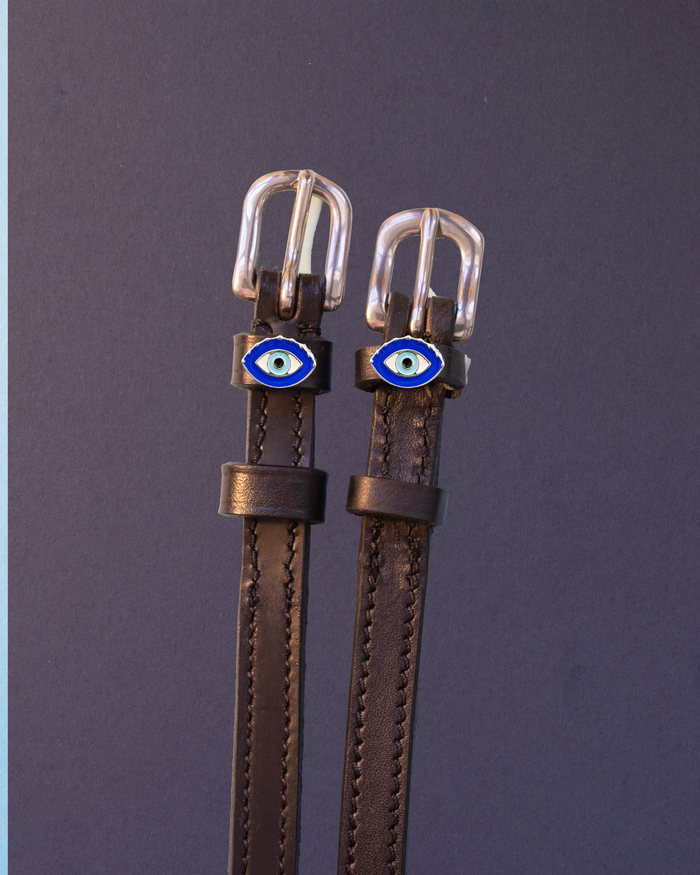 Brown leather evil eye ManeJane spur straps with silver buckles on a slate blue background