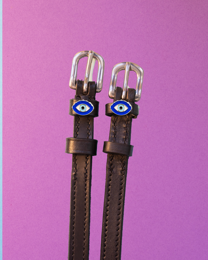 Brown leather ManeJane spur straps with blue evil eye charms on a purple background