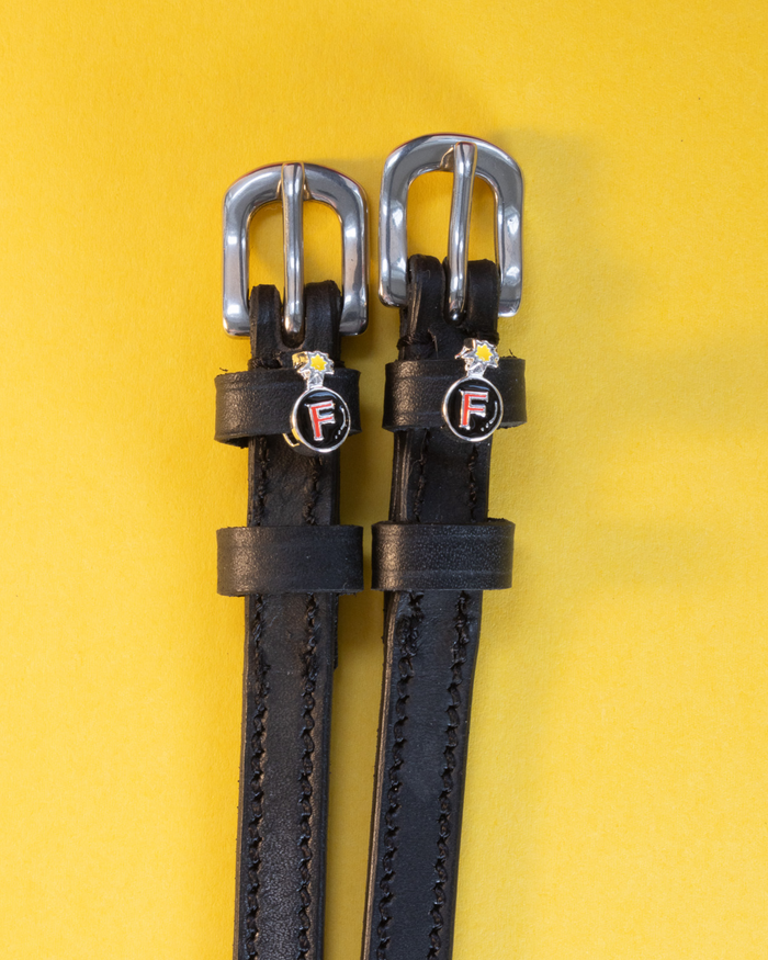 Pair of black leather F Bomb spur straps with silver buckles on a yellow background