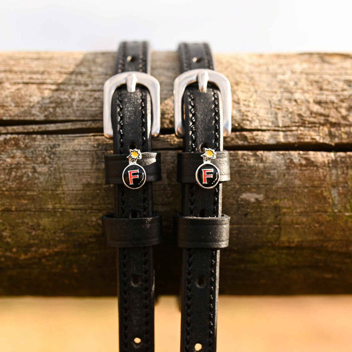 Black leather F Bomb spur straps hanging over a weathered wood rail with bomb charms