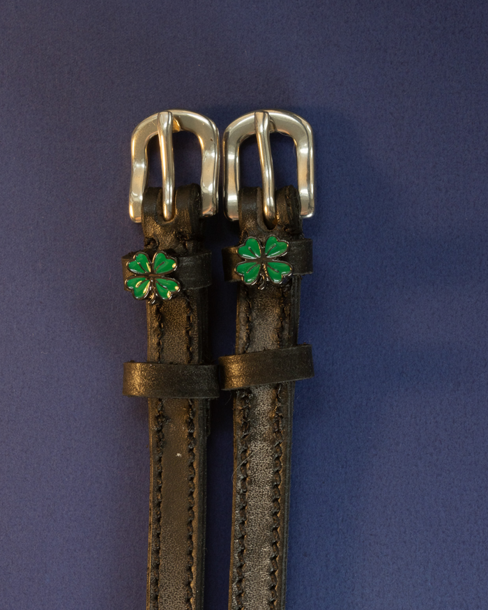 Black leather four leaf clover spur straps with green charms flat-laid on a navy background