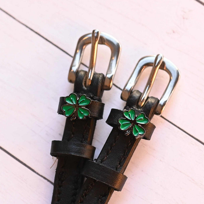 Angled close-up of ManeJane clover spur straps with green charms on pink wood planks