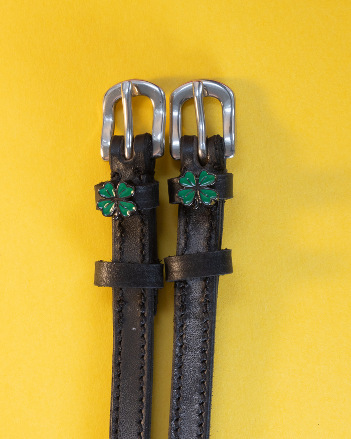 Black leather ManeJane spur straps with green four leaf clover charms on a yellow background