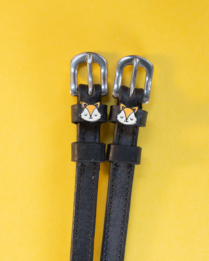 Black leather ManeJane spur straps with orange fox face charms on a yellow background