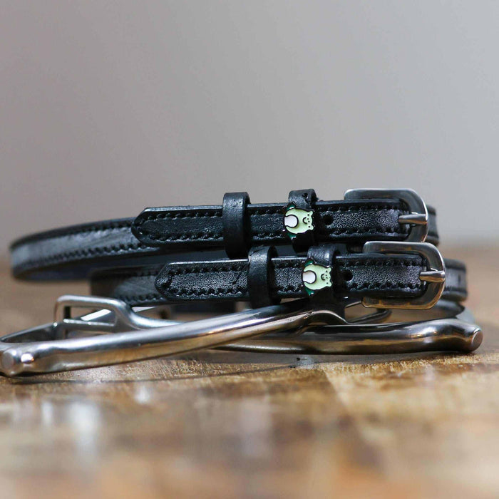 Black leather frog spur straps coiled beside a silver stirrup on a rustic wooden surface