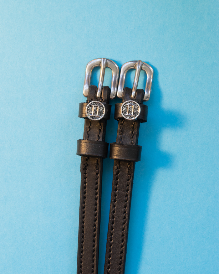 Black leather ManeJane Gemini spur straps with round zodiac charms on a light blue background